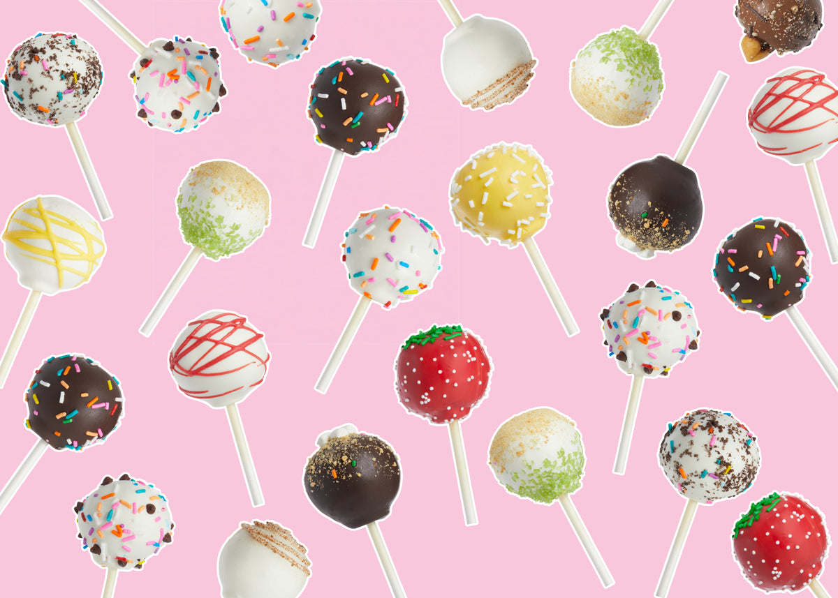 An assortment of decorated cake pops on white sticks, arranged in a repeating pattern on a solid light pink background. The cake pops are decorated with white, dark chocolate, yellow, green, red, and multicolor sprinkles.