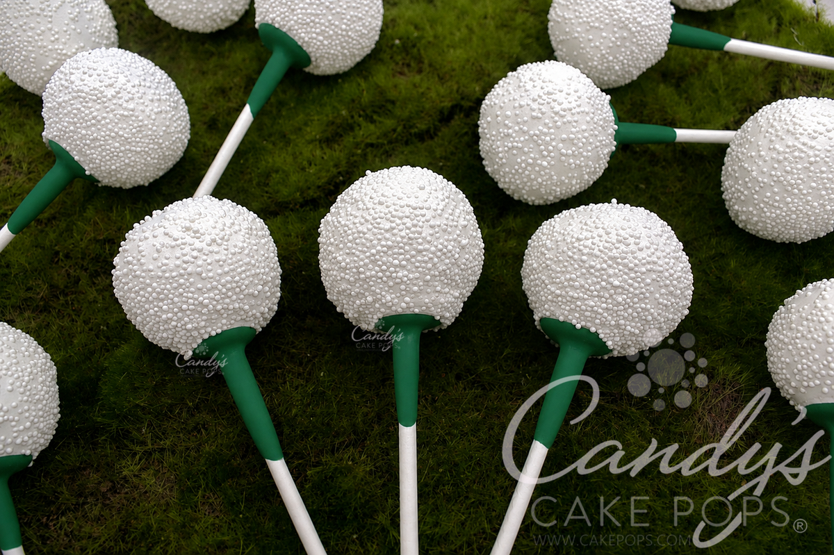 Golf Ball Cake Pops with green golf tees arranged on a green moss-like surface. For a Masters Golf Party/ A Candy's Cake Pops watermark is visible.