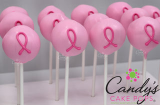 A close-up of several pink cake pops on white sticks, each decorated with a pink breast cancer awareness ribbon. The cake pops are arranged on a display stand against a light gray background, with a Candy's Cake Pops logo watermark in the lower right corner.