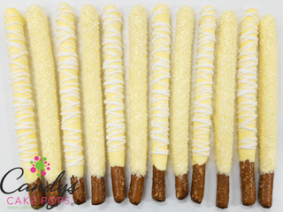 Twelve light yellow and white chocolate-covered pretzel sticks topped with white chocolate drizzle and sugar sprinkles, arranged upright on a white background. A Candy's Cake Pops logo is visible in the lower-left corner.