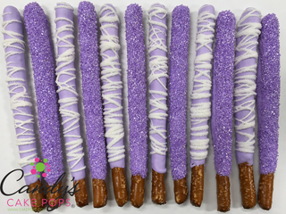 Twelve lavender and purple chocolate-covered pretzel sticks with white drizzle and sugar coating, arranged in two rows on a white background. A logo for Candy's Cake Pops is visible in the bottom-left corner.