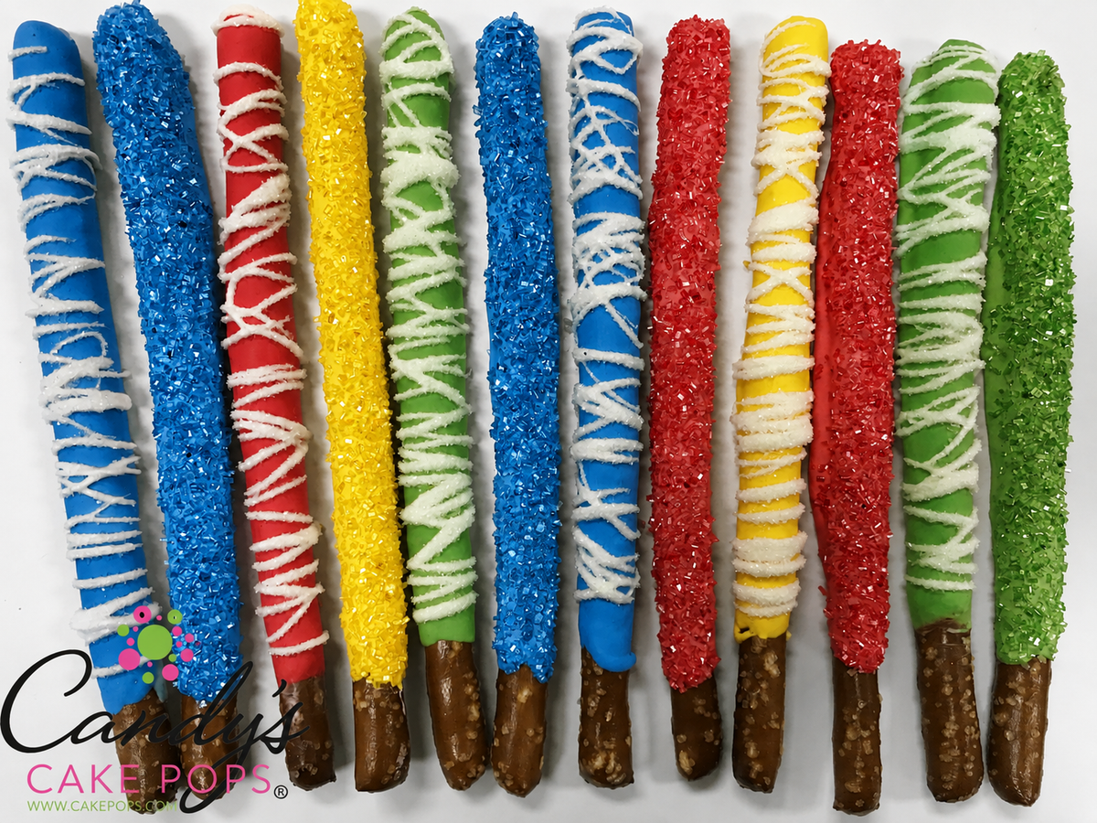 A row of colorful red, blue, yellow, and green chocolate-covered pretzel sticks decorated with glittery sugar sprinkles and white drizzle, displayed on a white background with a 'Candy's Cake Pops' logo in the lower left corner. Fun for a super hero party