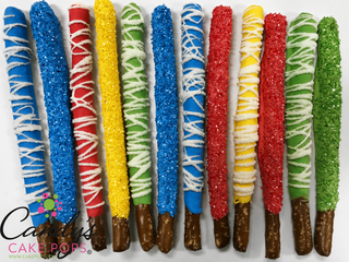 A row of colorful red, blue, yellow, and green chocolate-covered pretzel sticks decorated with glittery sugar sprinkles and white drizzle, displayed on a white background with a 'Candy's Cake Pops' logo in the lower left corner. Fun for a super hero party