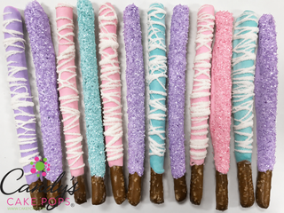 Twelve decorated chocolate-covered pretzel sticks arranged in a row on a white background. The pretzel tips are dipped in white chocolate and decorated with white drizzle, sugar crystals, or full purple, pink, and turquoise coatings and sprinkles